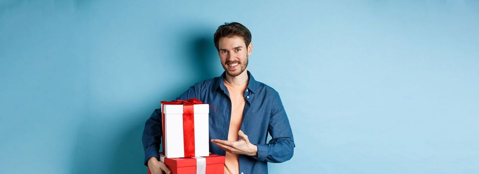 Young man in casual clothes buying romantic gifts on Valentines day, pointing at presents boxes and smiling, standing over blue background.