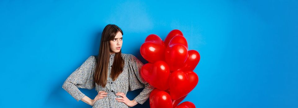 Valentines day. Annoyed and bothered girl complaining, looking left at empty space and frowning, standing near heart balloons, blue background.