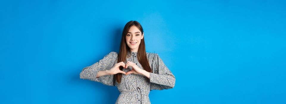 Pretty girl in romantic dress showing I love you heart gesture, smiling at camera, express sympathy and romance, standing on blue background.