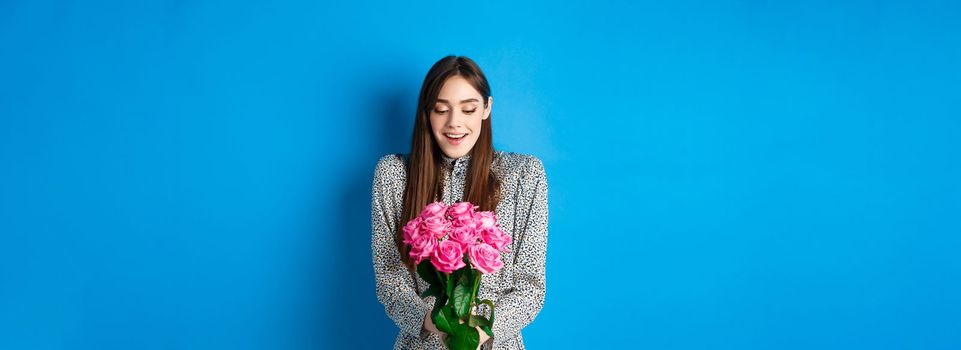 Valentines day concept. Happy attractive woman receive surprise flowers, looking thankful at bouquet of pink roses, standing on blue background.