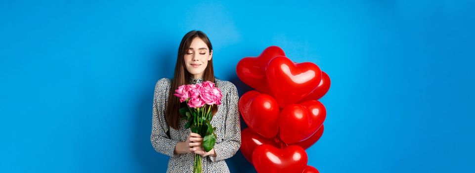 Valentines day. Romantic pretty woman close eyes and smelling beautiful flowers, standing near heart balloons, blue background.