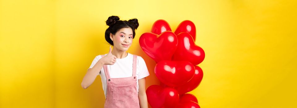Fashionble asian woman in romantic outfit with make-up, showing thumb up and smiling, praising Valentines day offer, standing near red heart balloons, yellow background.