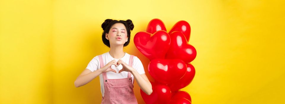 Romantic girl close eyes and pucker lips for kiss, showing I love you heart gesture, standing near cute red balloons, kissing lover over yellow background.