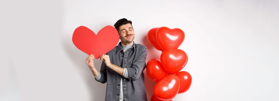 Romantic guy celebrating Valentines day, hugging big red heart card from lover and smiling happy, being in love, standing on white background near red balloons.