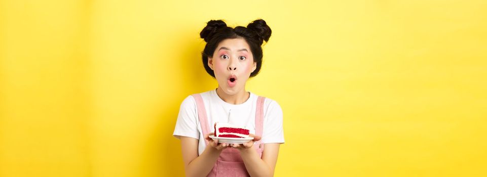 Holidays and celebration. Excited asian birthday girl blowing candle on party cake, making wish on b-day, standing against yellow background.