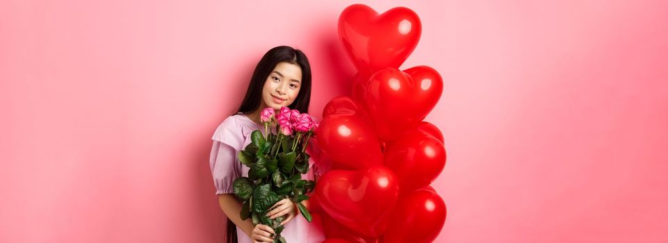 Valentines day concept. Tender and gentle teenage girl holding romantic flowers, standing near red heart balloons from lover and gazing with love, standing on pink background.
