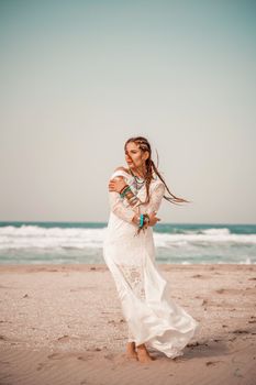 Model in boho style in a white long dress and silver jewelry on the beach. Her hair is braided, and there are many bracelets on her arms