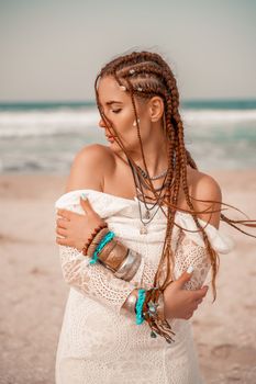 Model in boho style in a white long dress and silver jewelry on the beach. Her hair is braided, and there are many bracelets on her arms