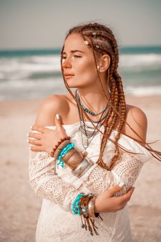 Model in boho style in a white long dress and silver jewelry on the beach. Her hair is braided, and there are many bracelets on her arms