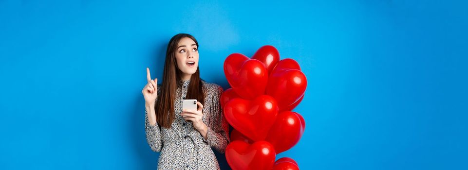 Valentines day. Image of romantic girl pitching an idea after using mobile phone, raising finger up and looking at empty space, standing near red heart balloons, blue background.