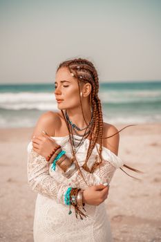 Model in boho style in a white long dress and silver jewelry on the beach. Her hair is braided, and there are many bracelets on her arms