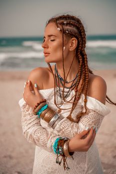 Model in boho style in a white long dress and silver jewelry on the beach. Her hair is braided, and there are many bracelets on her arms