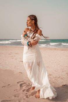 Model in boho style in a white long dress and silver jewelry on the beach. Her hair is braided, and there are many bracelets on her arms