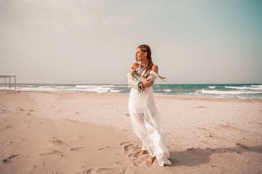 Model in boho style in a white long dress and silver jewelry on the beach. Her hair is braided, and there are many bracelets on her arms