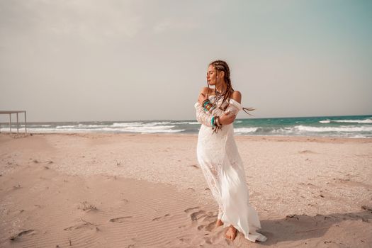 Model in boho style in a white long dress and silver jewelry on the beach. Her hair is braided, and there are many bracelets on her arms