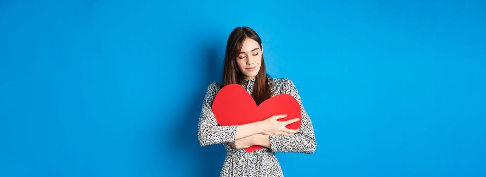 Valentines day. Romantinc girl in dress hugging big red heart cutout, close eyes and smile with dreamy face, imaging sensual date, standing on blue background.