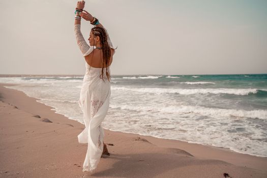 Model in boho style in a white long dress and silver jewelry on the beach. Her hair is braided, and there are many bracelets on her arms