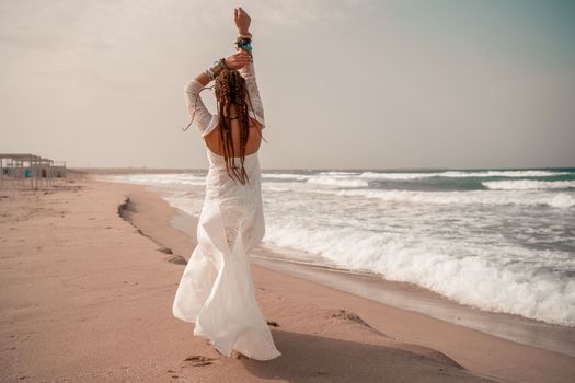 Model in boho style in a white long dress and silver jewelry on the beach. Her hair is braided, and there are many bracelets on her arms