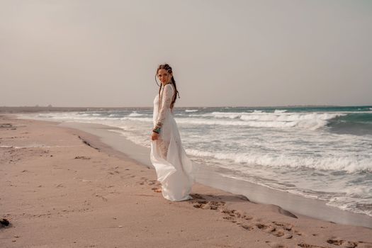 Model in boho style in a white long dress and silver jewelry on the beach. Her hair is braided, and there are many bracelets on her arms