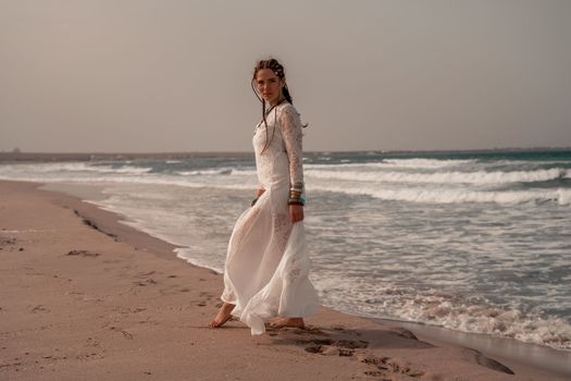 Model in boho style in a white long dress and silver jewelry on the beach. Her hair is braided, and there are many bracelets on her arms