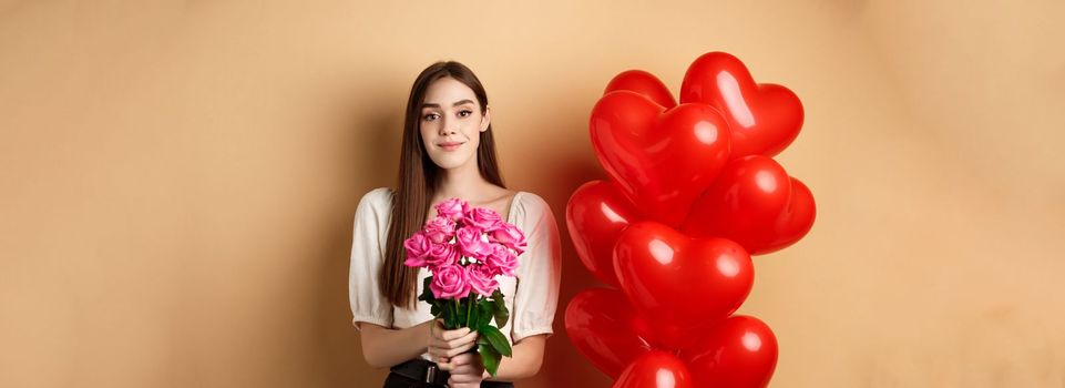 Beautiful girl holding bouquet of pink roses and smiling at camera, going on romantic date, standing near valentines heart balloons, beige background.