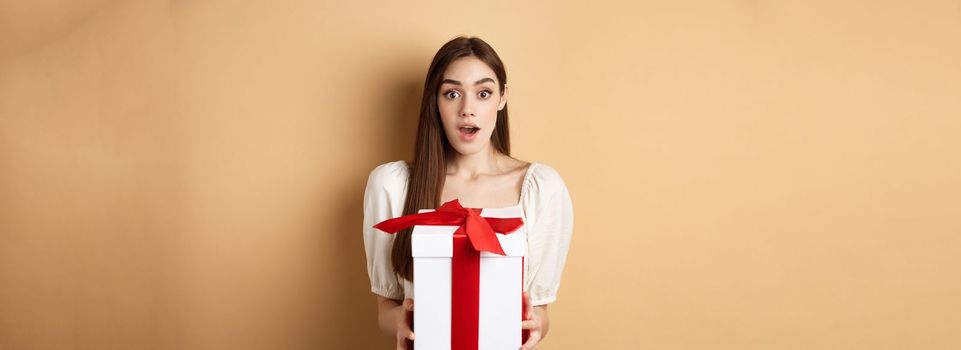 Amazed beautiful girl receiving gift from secret admirer on Valentines day, looking surprised at camera, holding box and standing on beige background.