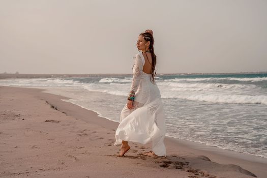 Model in boho style in a white long dress and silver jewelry on the beach. Her hair is braided, and there are many bracelets on her arms