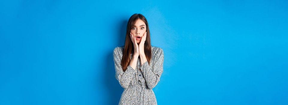 Shocked cute woman gasping amazed, drop jaw and stare with disbelief, standing in dress on blue background.