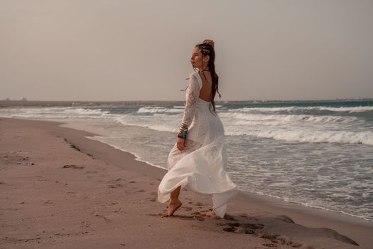 Model in boho style in a white long dress and silver jewelry on the beach. Her hair is braided, and there are many bracelets on her arms