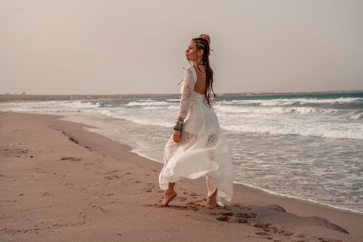 Model in boho style in a white long dress and silver jewelry on the beach. Her hair is braided, and there are many bracelets on her arms