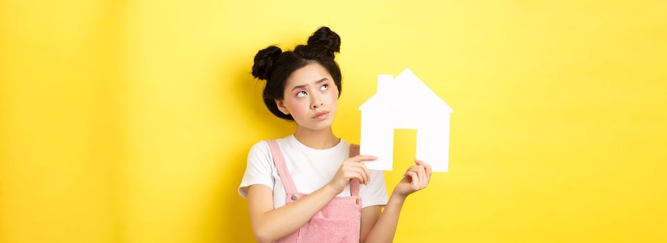 Real estate and family concept. Thoughtful asian girl dream of house, showing paper home cutout and look at upper left corner pensive, yellow background.