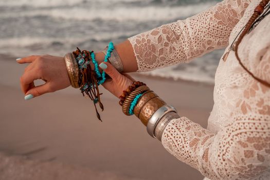 Model in boho style in a white long dress and silver jewelry on the beach. Her hair is braided, and there are many bracelets on her arms