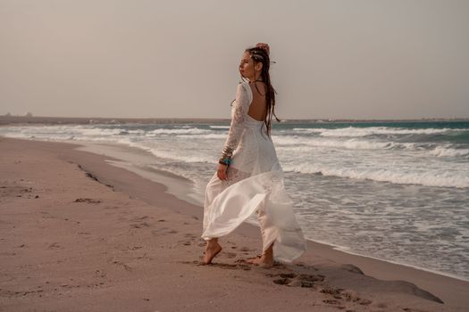 Model in boho style in a white long dress and silver jewelry on the beach. Her hair is braided, and there are many bracelets on her arms