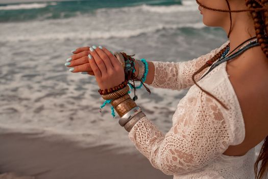 Model in boho style in a white long dress and silver jewelry on the beach. Her hair is braided, and there are many bracelets on her arms