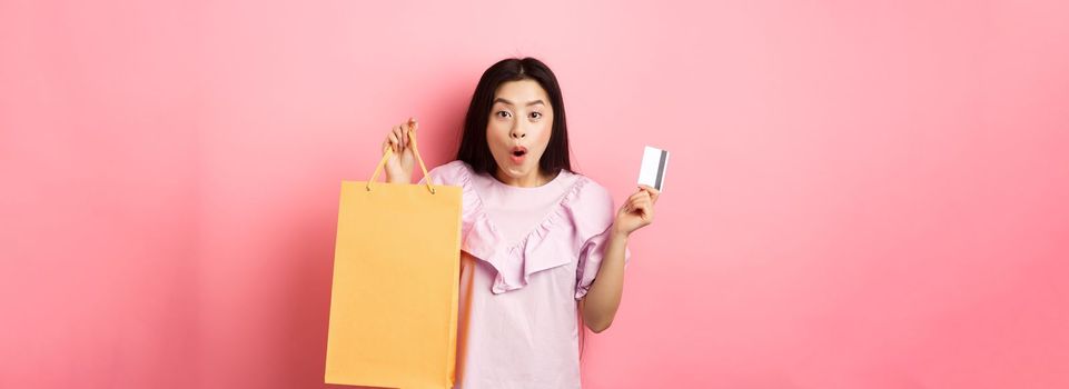 Shopping. Excited asian woman gasping amazed, holding bag from shop and plastic credit card, standing on pink background.