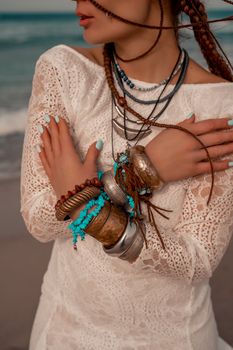 Model in boho style in a white long dress and silver jewelry on the beach. Her hair is braided, and there are many bracelets on her arms