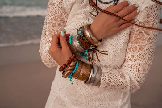 Model in boho style in a white long dress and silver jewelry on the beach. Her hair is braided, and there are many bracelets on her arms