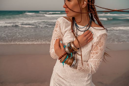 Model in boho style in a white long dress and silver jewelry on the beach. Her hair is braided, and there are many bracelets on her arms