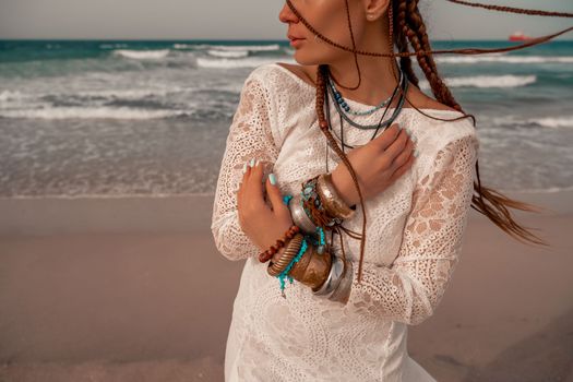 Model in boho style in a white long dress and silver jewelry on the beach. Her hair is braided, and there are many bracelets on her arms