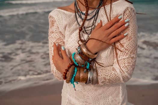 Model in boho style in a white long dress and silver jewelry on the beach. Her hair is braided, and there are many bracelets on her arms