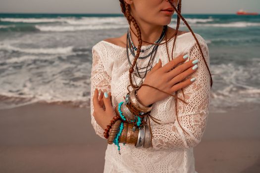 Model in boho style in a white long dress and silver jewelry on the beach. Her hair is braided, and there are many bracelets on her arms