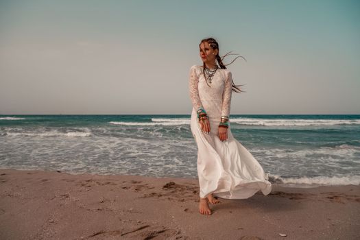 Model in boho style in a white long dress and silver jewelry on the beach. Her hair is braided, and there are many bracelets on her arms