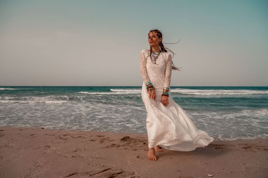 Model in boho style in a white long dress and silver jewelry on the beach. Her hair is braided, and there are many bracelets on her arms