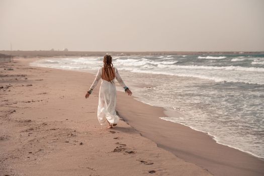 Model in boho style in a white long dress and silver jewelry on the beach. Her hair is braided, and there are many bracelets on her arms