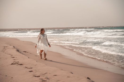 Model in boho style in a white long dress and silver jewelry on the beach. Her hair is braided, and there are many bracelets on her arms