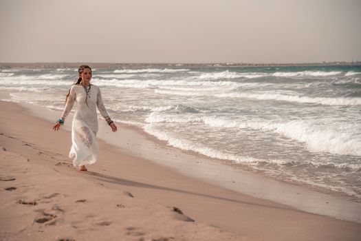 Model in boho style in a white long dress and silver jewelry on the beach. Her hair is braided, and there are many bracelets on her arms