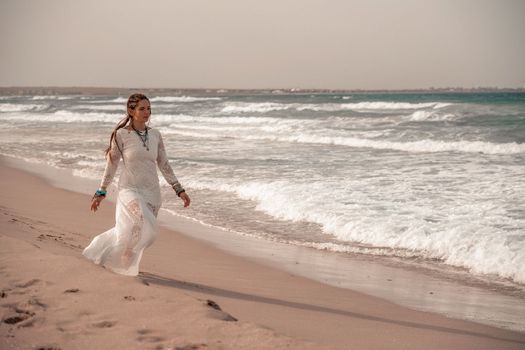 Model in boho style in a white long dress and silver jewelry on the beach. Her hair is braided, and there are many bracelets on her arms