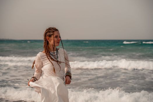 Model in boho style in a white long dress and silver jewelry on the beach. Her hair is braided, and there are many bracelets on her arms