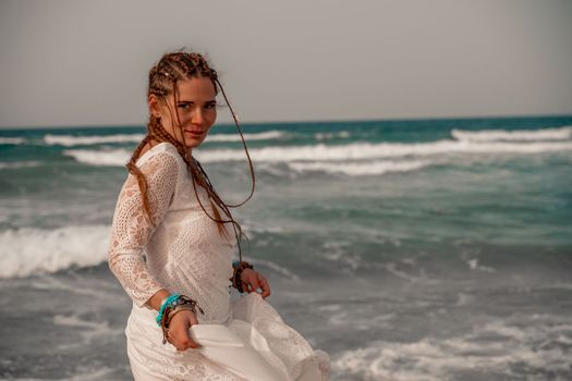 Model in boho style in a white long dress and silver jewelry on the beach. Her hair is braided, and there are many bracelets on her arms