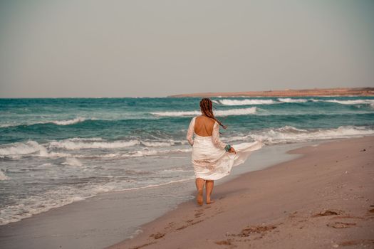 Model in boho style in a white long dress and silver jewelry on the beach. Her hair is braided, and there are many bracelets on her arms